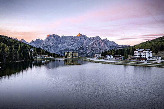 View of the serene Lake Misurina reflecting the majestic Dolomites under a pastel dawn sky, framed by evergreen forests, Campitello di Fassa, Trentino-Alto Adige, Italy.