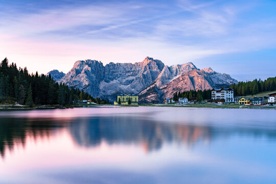 View of the tranquil lake mirroring the towering Dolomites under a serene sky painted with pastel hues, fringed by lush forests, Campitello di Fassa, Trentino-Alto Adige, Italy.