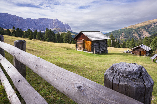 View of wooden cabins nestled in a vibrant green meadow, framed by a rustic wooden fence against a backdrop of majestic mountains, Campitello di Fassa, Trentino-Alto Adige, Italy.