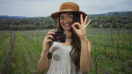 Obraz premium Young woman smiling face holds dslr camera and makes ok gesture in lush forest under green vines; confidence.