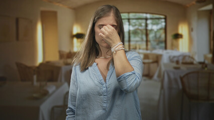 Woman in blue shirt pinching nose at restaurant table indoors under bright ambient lighting; disgust.
