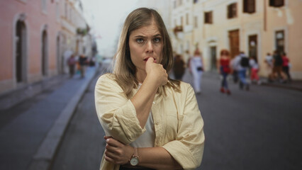 Fototapeta premium Blonde woman wearing a beige shirt biting her finger on a pedestrian street in front of historic buildings; anxiety.