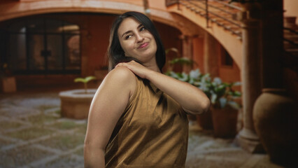 Fototapeta premium Young hispanic woman rubbing her bare shoulder at building entrance in warm sunlight, casual brown top and relaxed pose; contentment.