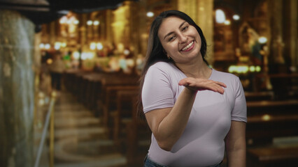 Fototapeta premium Young woman smiling and blowing a kiss with open palms and visible hands inside a catholic church aisle lit by lamps; joy faith devotion.