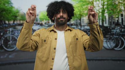 Man with crossed fingers on a street in amsterdam, curly hair and raised hands beside canal...