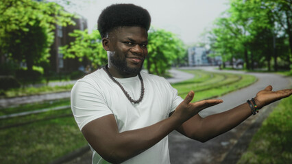 Obraz premium Man showing thumbs up and presenting open palms in street park wearing white t shirt and beaded necklace; confidence welcome.