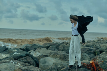 Female on a rocky shoreline facing strong wind with flowing coat, casual chic fashion scene by the...