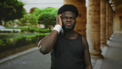 Fototapeta premium Man talking on smartphone, wristband on forearm, showing bicep in building colonnade beside a green park path; concern solitude.