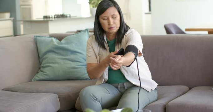 Mature Asian woman sitting on couch, wrapping BP cuff and adjusting monitor to check BP