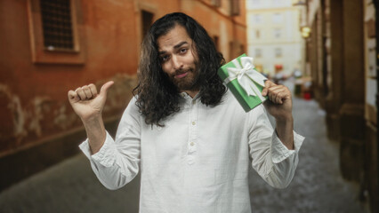 Man pointing thumb to chest while holding a wrapped green gift box with white bow in a cobbled street between old buildings; self promotion confidence.
