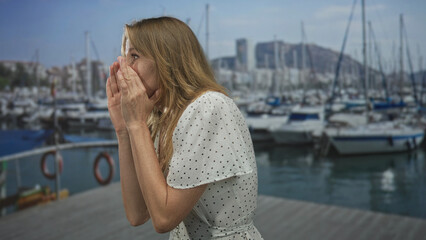 Fototapeta premium Woman calling with cupped hands and open arms on marina dock beside moored boats in seaside port; urgent call.