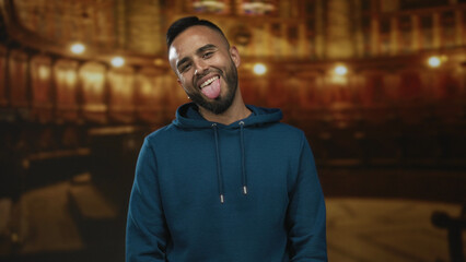 Man sticking tongue out and leaning forward in a teal hoodie inside an ornate church building with blurred pews and warm lights  playful mischief casual defiance. © Krakenimages.com