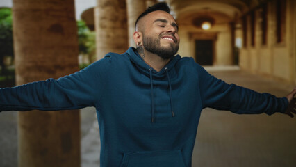 Man wearing teal hoodie with arms outstretched in old town building corridor, smiling with closed...