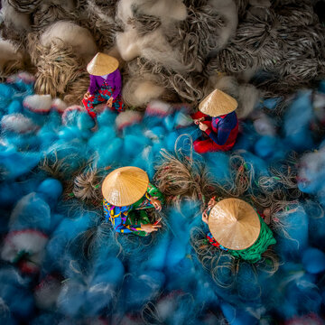 View of women in colorful clothes and conical hats mending a sea of vibrant blue fishing nets and piles of soft white cotton, Bac Lieu, Mekong Delta, Vietnam.