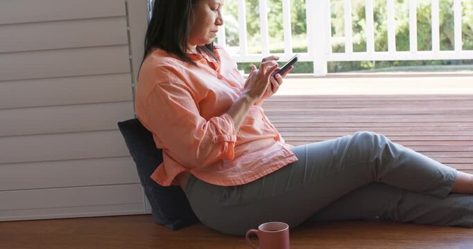 Mature female sitting on porch floor against navy cushion scrolling phone with pink mug nearby
