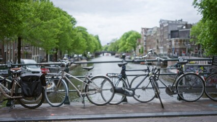 Fototapeta premium Bicycles parked on a bridge in amsterdam with a blurred view of canals and historic buildings under a cloudy sky captured in a defocused aesthetic.