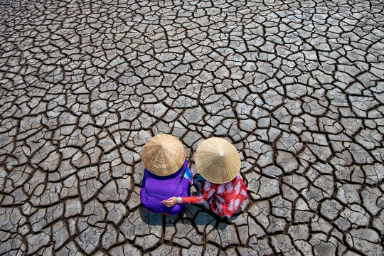 View of two women in conical hats crouch on parched earth, the cracked soil a stark tapestry of drought, in a poignant tableau, Bac Lieu, Mekong Delta, Vietnam.