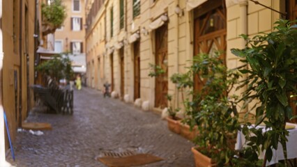 Obraz premium Sunlit narrow cobblestone alley with blurred storefronts, stacked chairs and potted plants in soft bokeh alley; background backdrop copyspace backplate.