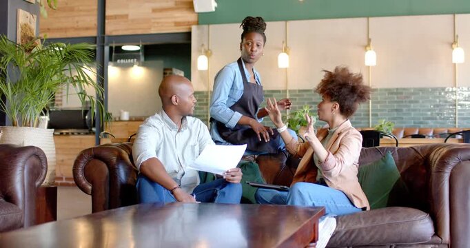 African American server placing cup on sofa while clients in cafe reading papers tablet man looking