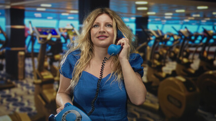 Woman holding blue rotary telephone to ear in studio gym scene with treadmills and machines blurred...