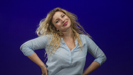 Fototapeta premium Woman with hands on hips in blue studio, smiling toward camera with tousled blonde hair and light blouse; confidence vitality.