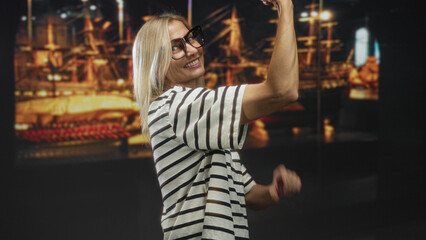 Fototapeta premium Woman flexing arm toward ship model in building, wearing striped shirt and large glasses while smiling in side profile; confidence celebration.