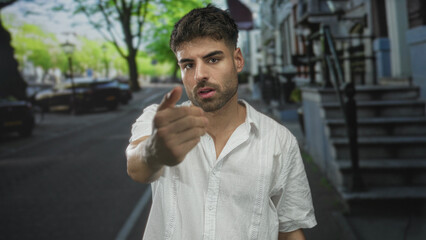 Man beckoning finger on tree lined street with blurred parked cars and black wrought iron fences in...