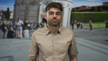 Young hispanic man wearing glasses and casual shirt shrugs beside leaning tower building with puzzled expression; uncertainty.