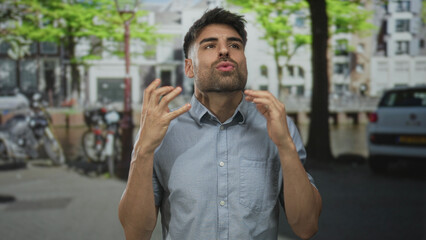 Fototapeta premium Man raising hands by canal on bustling street lined with parked bikes and cars under green trees; frustration.