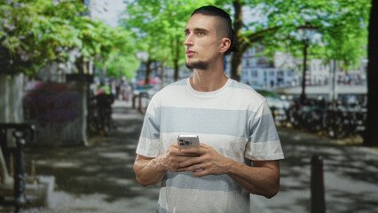 Fototapeta premium Man holding smartphone with both hands and looking aside on a tree lined city street with bicycles and parked cars; contemplation urban life.