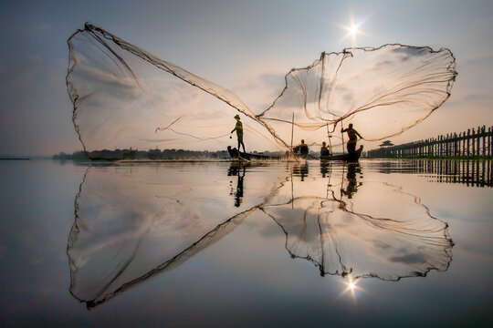 View of fishermen casting nets that are reflected in the still waters near a bridge at sunrise, U Bein Bridge, Mandalay, Myanmar.
