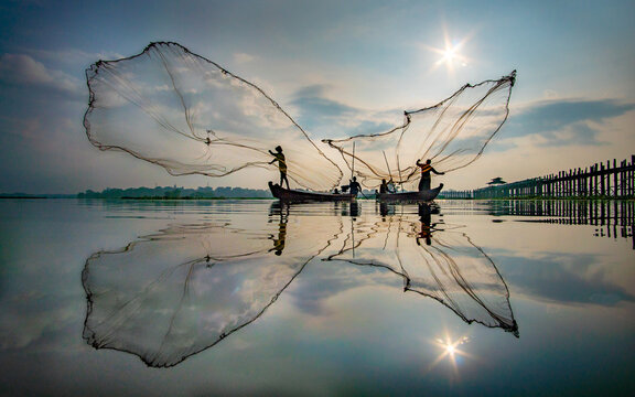View of fishermen casting nets from their boats, the mirrored expanse reflecting the tranquil sky, near the U Bein Bridge, Mandalay, Myanmar.