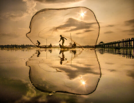 View of silhouette fishermen casting nets from wooden boats on a serene lake with the sun reflecting in the water, U Bein Bridge, Mandalay, Myanmar.