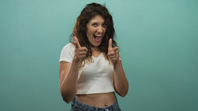 Young woman with curly hair smiling and pointing finger guns toward camera in teal studio wearing a cropped white top and jeans; playful confidence.