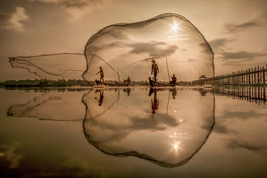 View of fishermen casting their nets from wooden boats on calm waters reflecting the golden sunset and U Bein Bridge, Mandalay, Myanmar.