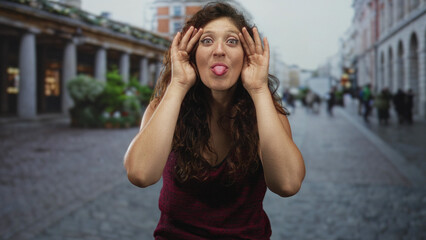 Fototapeta premium Young hispanic woman laughing with hands to face and covering eyes on a crowded cobbled street beside a colonnade of shops; joy playful surprise.