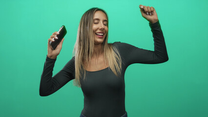 Young woman smiling and dancing while holding smartphone with arms raised in bright green studio;...
