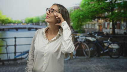 Fototapeta premium Woman tucks hair behind ear while adjusting glasses on canal street in amsterdam, near bikes and water; calm reflection.
