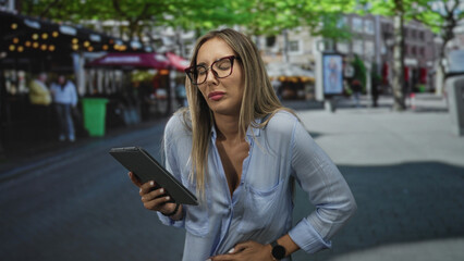 Fototapeta premium Woman holding tablet, hand on lower back for backache on street near cafes and trees; discomfort busy day.