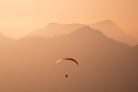 View of a lone paraglider soars through the hazy, orange-tinted sky, with layered silhouettes of mountains fading into the distance, Riemenstalden, Schwyz, Switzerland.