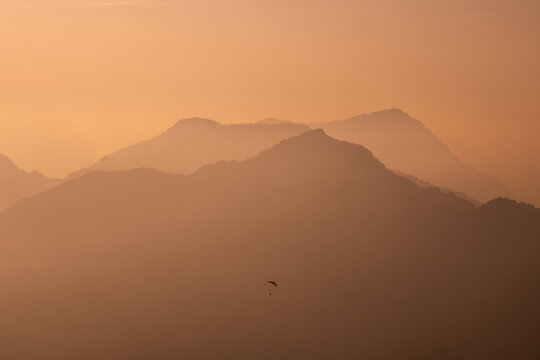 View of a lone paraglider soaring amidst the hazy, sun-kissed mountain silhouettes, painted in warm amber tones, Riemenstalden, Schwyz, Switzerland.