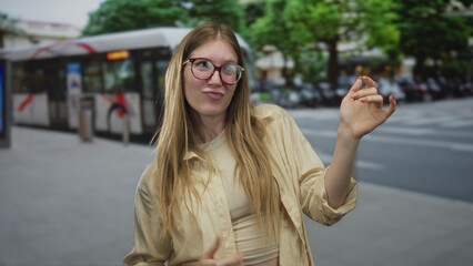 Fototapeta premium Young blonde woman wearing glasses performs air guitar hand gesture on city street in front of a bus; joy.