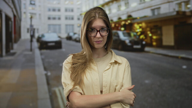 Blonde woman in glasses yawns with hand on neck on street under soft evening lights amid blurred cars; fatigue.