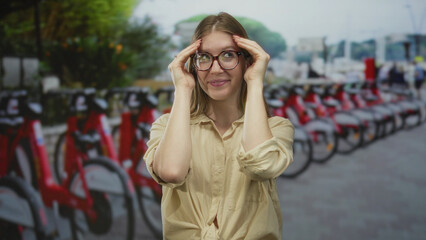 Obraz premium Woman wearing glasses presses temples beside red rental bikes lined in a row on busy city street; relief calm.