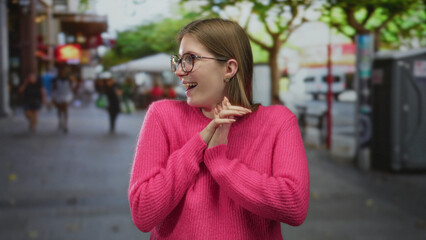 Fototapeta premium Young blonde woman smiling broadly and showing open palms on a busy city street lined with trees and cafes; joy.