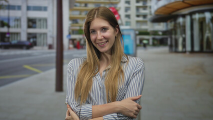 Fototapeta premium Woman with arms crossed on city street in striped shirt near modern storefronts, smiling softly at camera; confidence and ease.