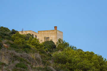 Fototapeta premium A view of a solitary, earth-toned building perched on a hillcrest, set against a clear blue sky and surrounded by dense Mediterranean scrub and pine trees, symbolizing remote island living and escape.