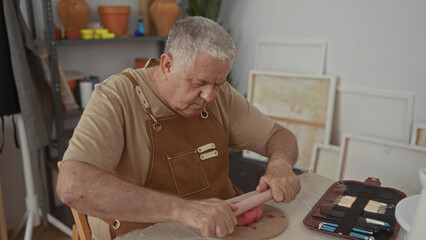 Man in apron presses and rolls clay with hands and rolling pin beside pottery tools on table in...
