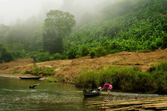 View of a woman in vibrant pink, working by a boat near the lush riverbank under a hazy sky, creating a serene yet industrious scene, Bandarban, Bangladesh.