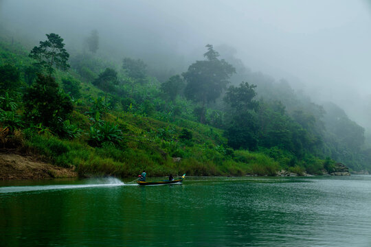 View of a small boat slicing through the emerald river beneath a mist-shrouded, verdant hillside in Tindu village, Bandarban, Chittagong Division, Bangladesh.